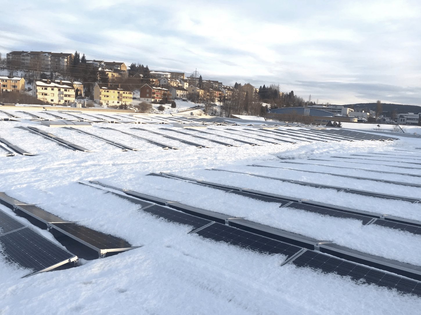 Snødekte solcellepaneler i et boligområde med hus i bakgrunnen