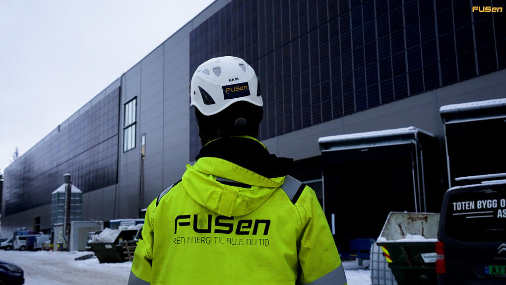 FUSen-ansatt i gult arbeidsutstyr foran en bygning med installerte solcellepaneler på fasaden, vinterdag