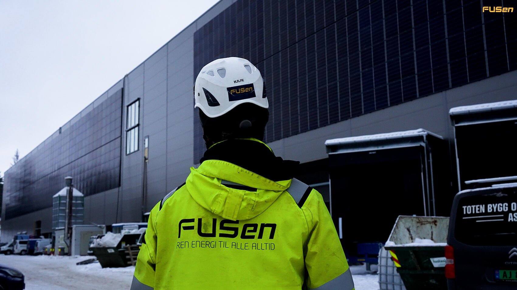 FUSen-ansatt i gult arbeidsutstyr foran en bygning med installerte solcellepaneler på fasaden, vinterdag.